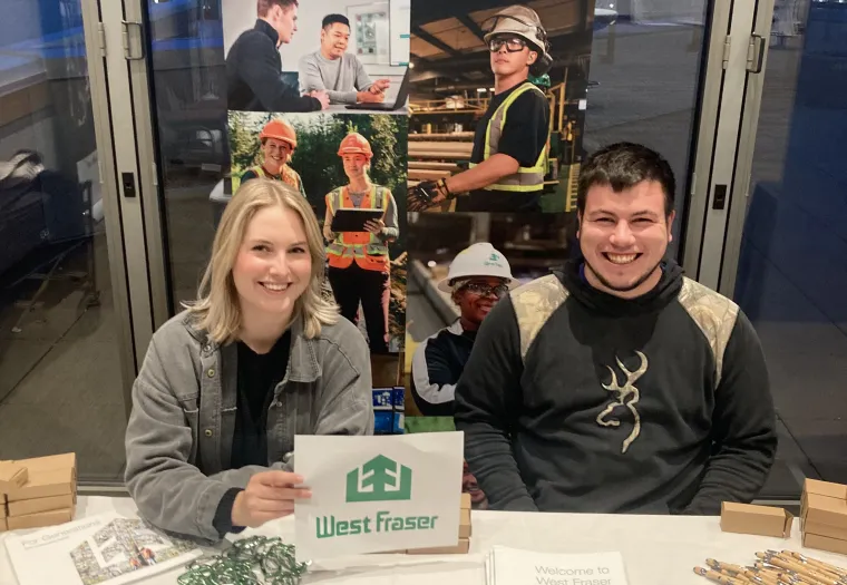 Two people sit at a recruitment table for West Fraser, displaying informational materials and promotional items in front of a "Build Your Career" banner.