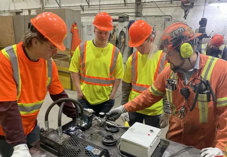 Three students and one worker in safety gear examine machinery components on a table in an industrial workshop setting.