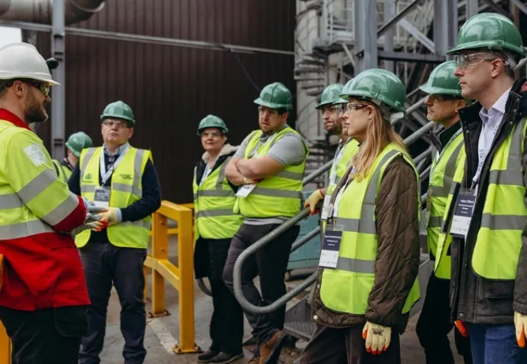 A group of people wearing hard hats, safety vests, and gloves listen to a worker in a red jacket at an industrial site.