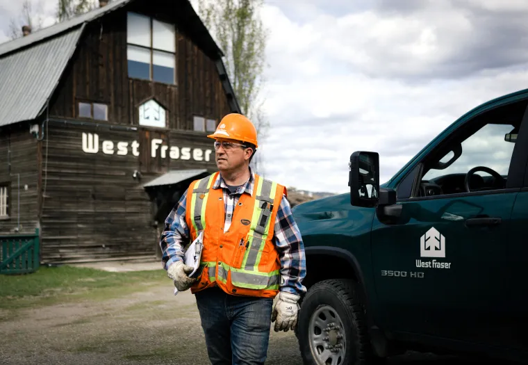 A man in a safety vest and hard hat walks near a West Fraser building and company truck on a cloudy day.