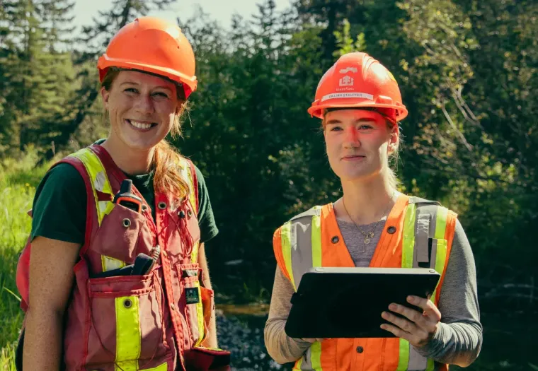 Two people wearing safety helmets and reflective vests stand outdoors; one is smiling and the other holds a tablet. Trees and greenery are visible in the background.