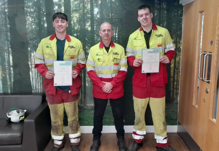 Three people in safety uniforms stand indoors; two hold certificates and one stands in the center. The background shows a forest wall mural and part of a wooden door.