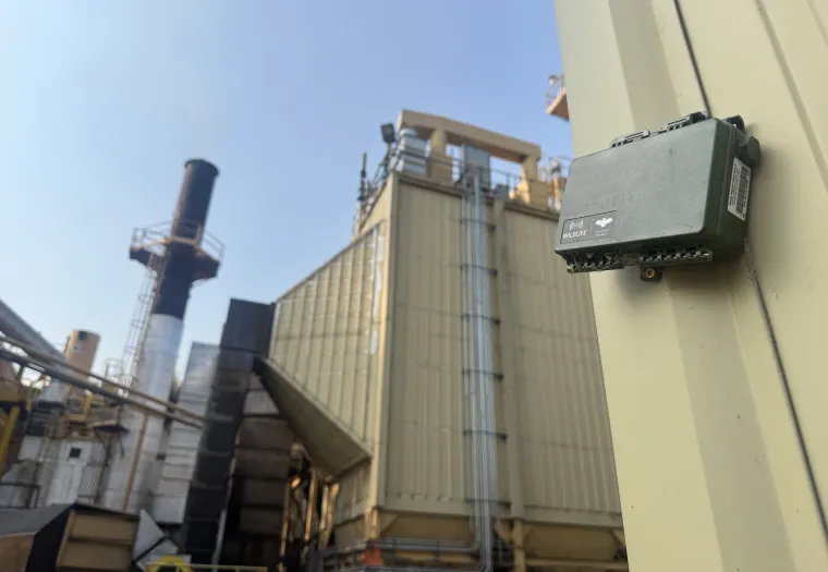 A gray Wildlife Acoustics audio recorder mounted on a metal industrial structure at a facility with smokestacks.
