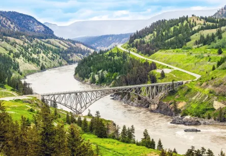 A steel bridge spans a wide, winding river in a mountainous, forested landscape of Williams Lake. The scene includes a curving road and patches of green hills under a partly cloudy sky.