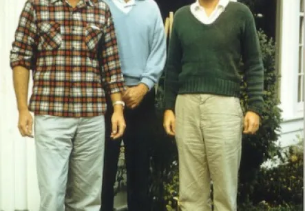Three men standing in front of a white building with a brick step, dressed in casual clothing: one in plaid, one in a light blue sweater, and one in a green sweater.