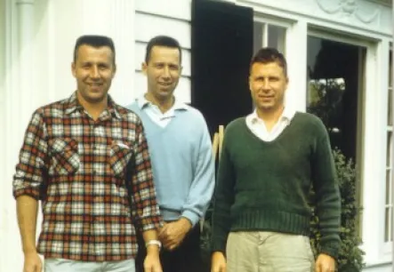 Three men standing in front of a white building with a brick step, dressed in casual clothing: one in plaid, one in a light blue sweater, and one in a green sweater.
