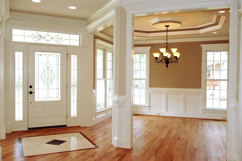 A bright entryway with a white front door, sidelights, large windows, hardwood floors, and a view into a dining room with a chandelier and coffered ceiling.