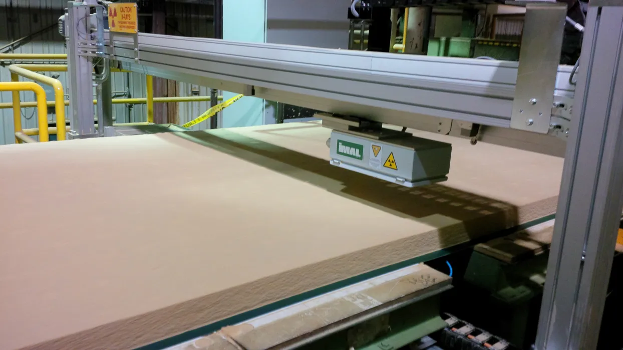 A machine processes a large sheet of material, possibly wood or fiberboard, in an industrial setting with safety signs visible.