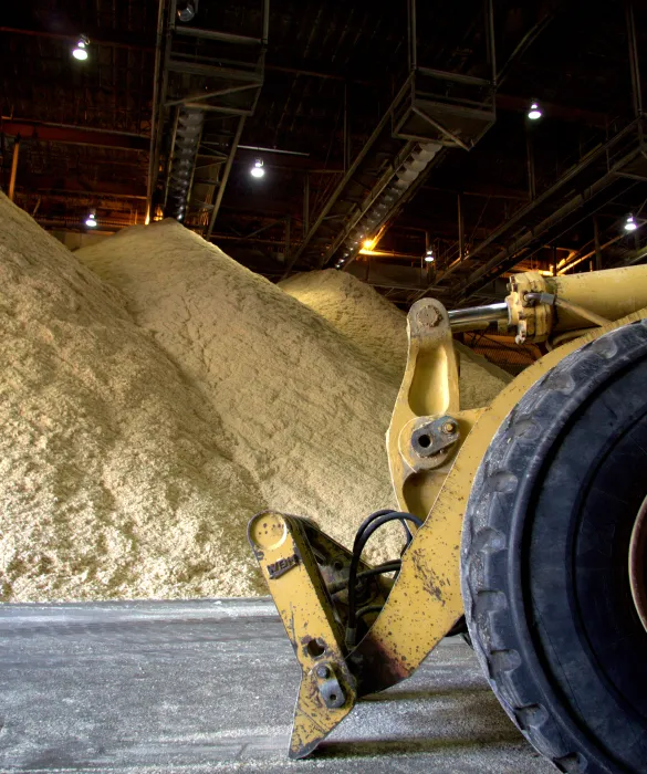 A large yellow front-end loader is parked inside an industrial warehouse with tall piles of sawdust or wood chips in the background.