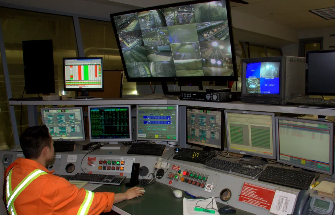 Person in a high-visibility jacket sits at a control room desk, monitoring multiple computer screens and security camera feeds.