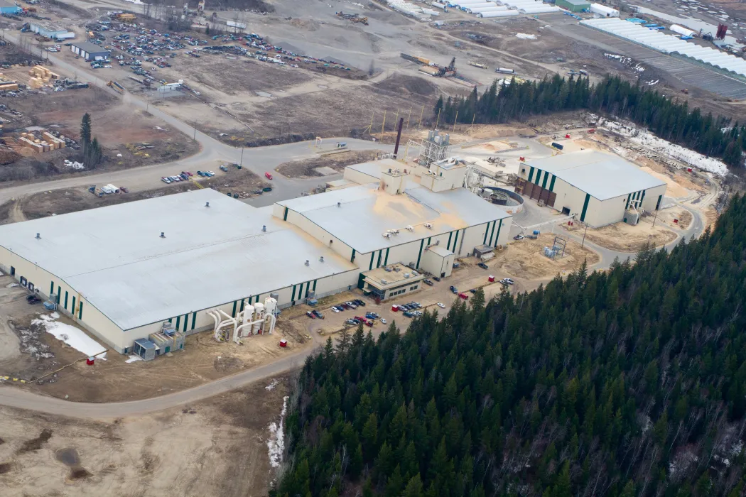 Aerial view of a large industrial facility surrounded by a forested area and dirt roads, with parked vehicles and smaller structures nearby.
