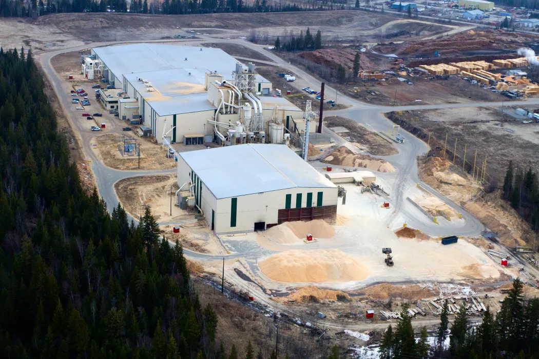 Aerial view of an industrial facility with large buildings, wood processing equipment, and piles of sawdust, surrounded by forest and dirt roads.