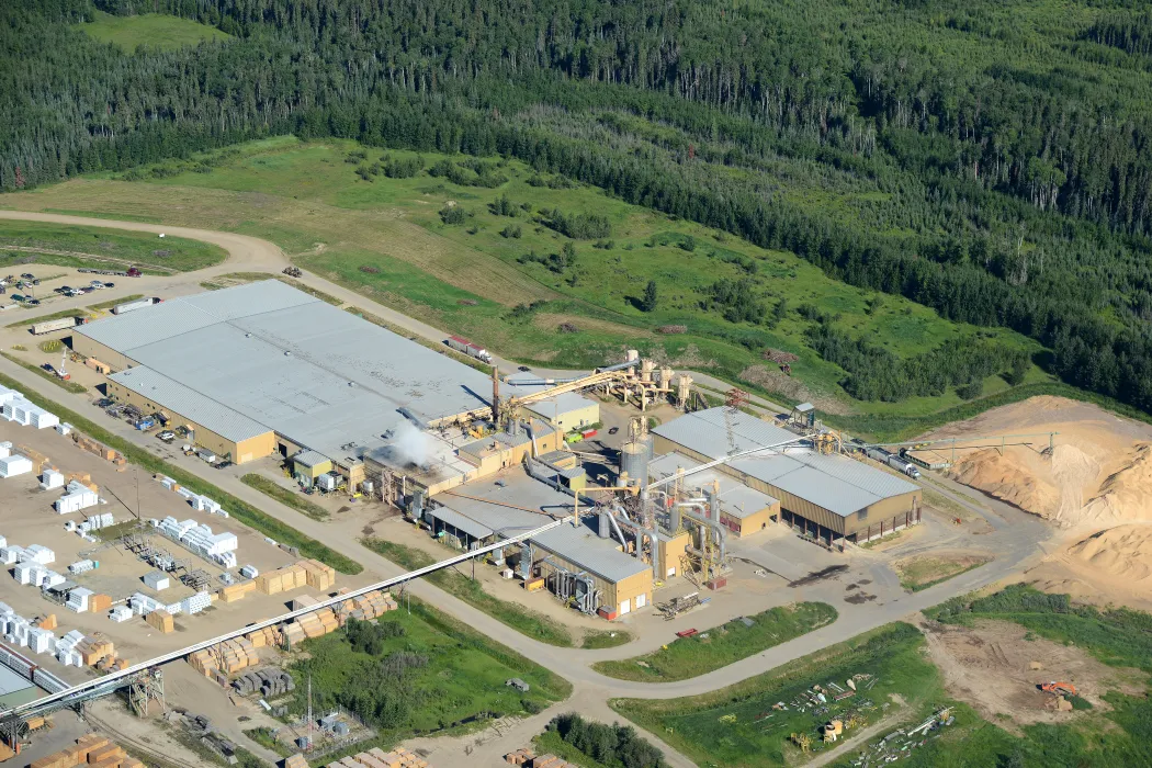 Aerial view of an industrial facility surrounded by forest, with large buildings, outdoor storage, piles of materials, and some visible machinery.