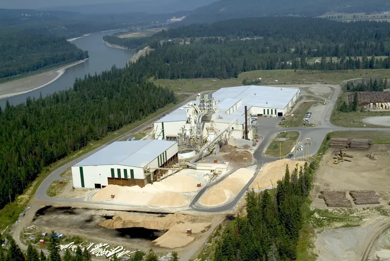 Aerial view of a lumber mill facility surrounded by forest, with wood piles, buildings, and a river in the background.