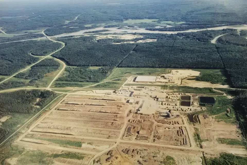 Aerial view of an industrial facility with cleared land, buildings, storage piles, and surrounding forested area. Roads and a river are visible in the background.