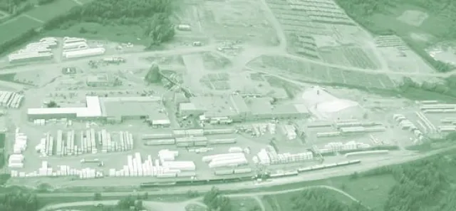 Aerial view of an industrial complex surrounded by wooded areas, showing large buildings, stacks of materials, and trucks on-site.