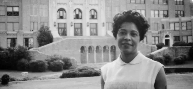 A woman is standing in front of a large building with multiple windows and a grand staircase. She is wearing a sleeveless shirt and smiling at the camera.