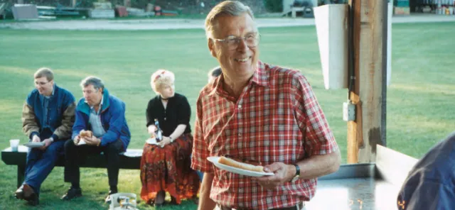 A man in a red checkered shirt holds a hot dog on a plate while standing outdoors at a social gathering. Three people are seated in the background.