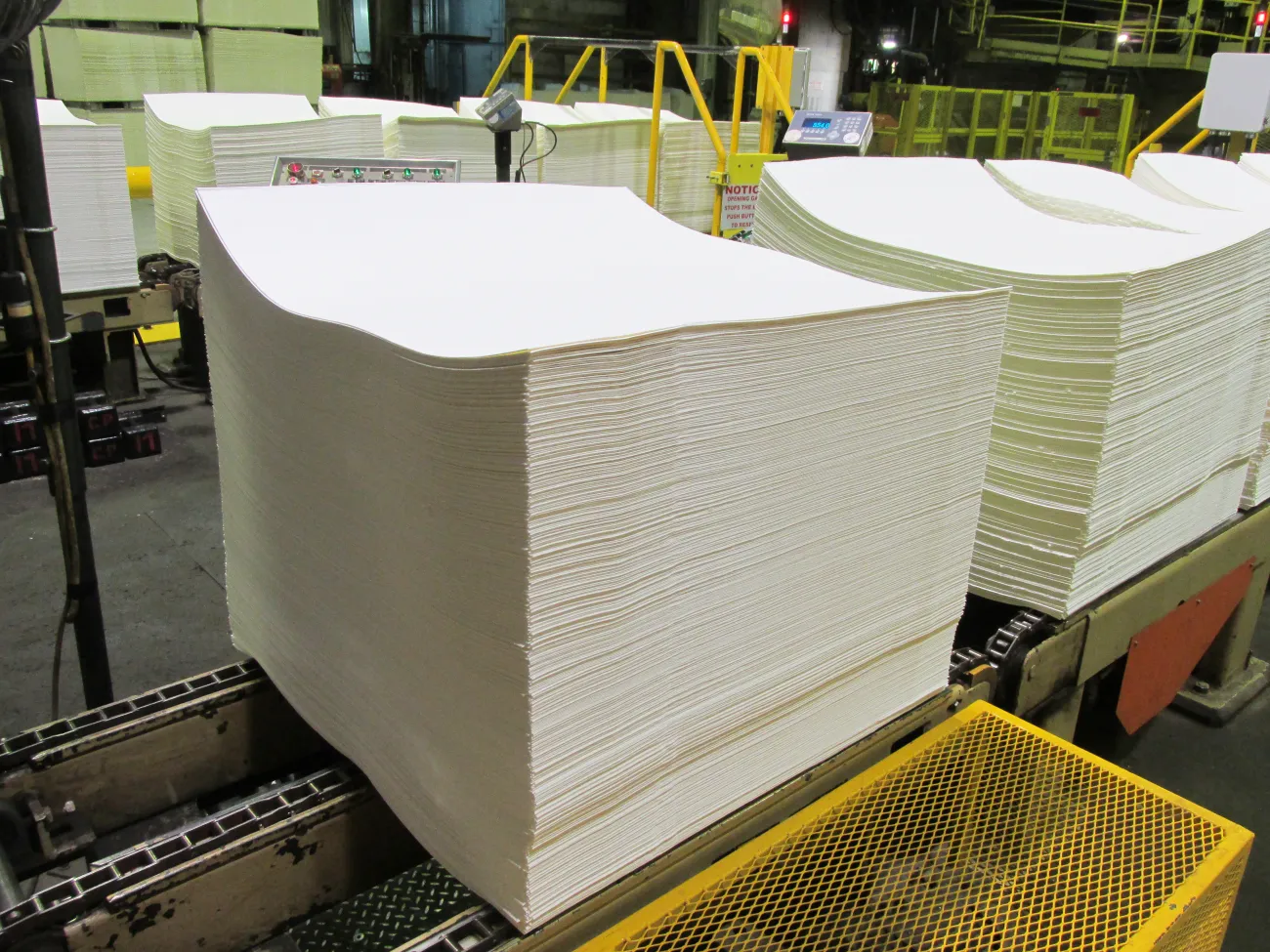 A large stack of blank white paper sheets on an industrial conveyor belt inside a factory setting.