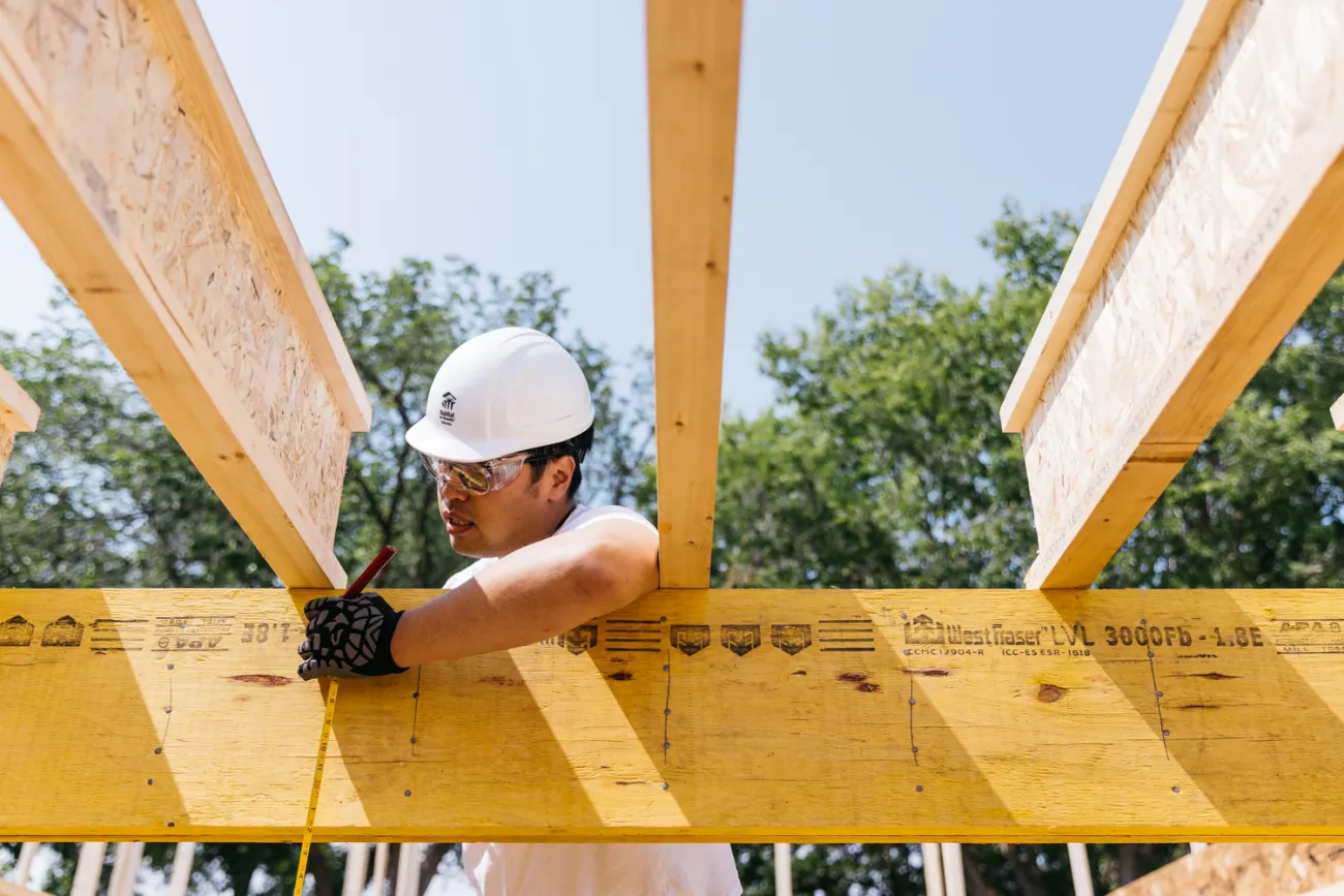 A construction worker wearing a white hard hat and gloves marks measurements on a wood beam using a pencil at a construction site.