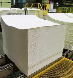 A large stack of blank white paper sheets on an industrial conveyor belt inside a factory setting.