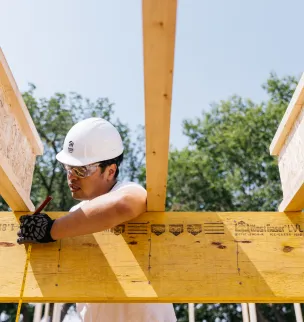 A construction worker wearing a white hard hat and gloves marks measurements on a wood beam using a pencil at a construction site.