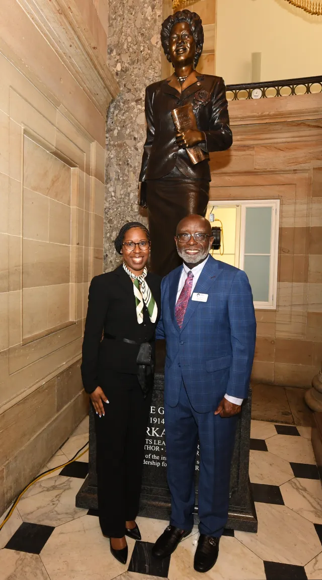 Two people stand in front of a statue in a well-lit, ornate hallway. The individual on the left is dressed in a black suit with a patterned scarf, and the one on the right is dressed in a blue suit.
