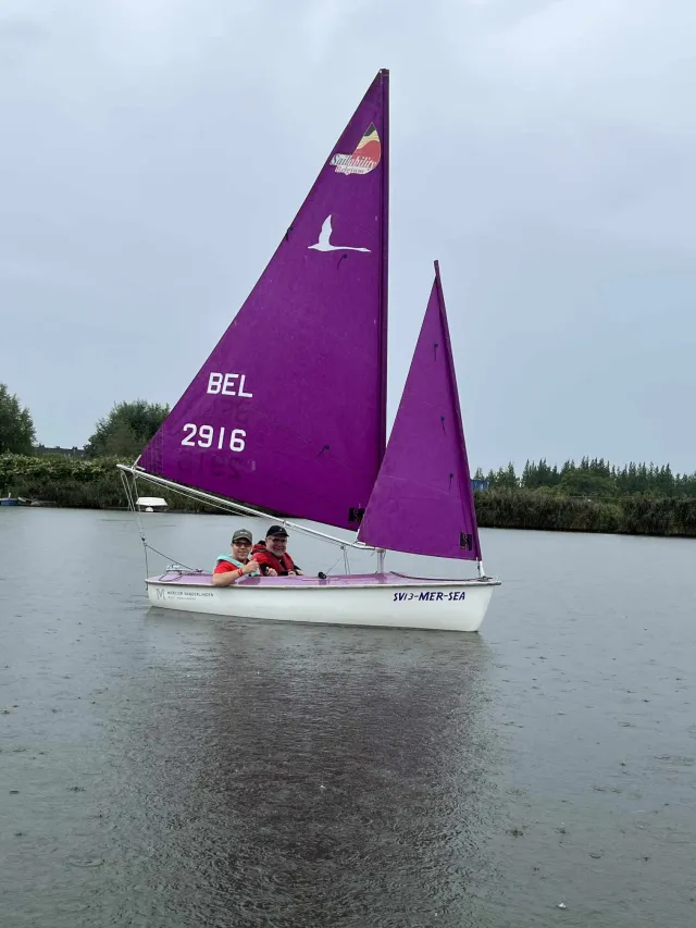 Two people participate in non-profit Vilvoorde, Belgium-based sailing club WSKLuM's adaptive sailing program. They sail a small white boat with a bright purple sail on a calm body of water, surrounded by trees under a cloudy sky.