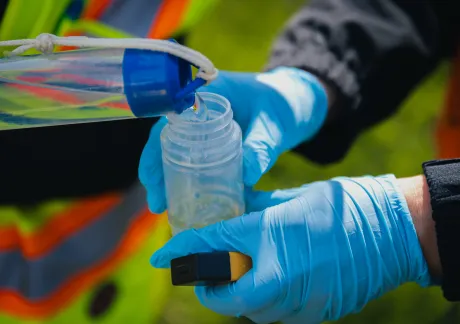 Two gloved hands transfer liquid from a container into a smaller bottle outdoors.