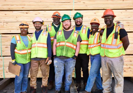 A group of seven construction workers wearing safety helmets and reflective vests stand together in front of a stack of wooden planks.
