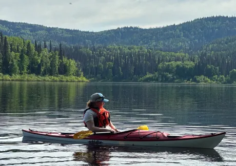 A person in a red and white kayak floats on a calm lake surrounded by lush, green forested hills. The individual wears a life vest and hat.