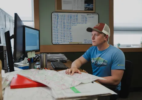 A West Fraser worker wearing a cap and a blue shirt sits at a desk with a computer, keyboard, and maps, surrounded by office materials and a whiteboard in the background.