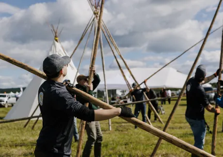 Several people work together to set up wooden poles for a large tent structure or teepee frame in an open field on a cloudy day.