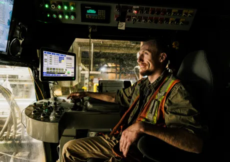 Person in a high-visibility vest operates machinery, sitting in a control room with multiple screens and control panels.