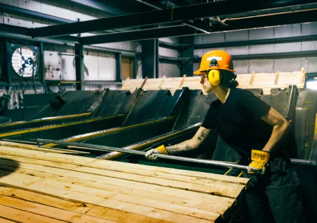 A worker wearing protective gear handles wooden planks in an industrial setting, with machinery visible in the background.