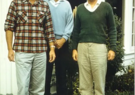 Three men standing in front of a white building with a brick step, dressed in casual clothing: one in plaid, one in a light blue sweater, and one in a green sweater.