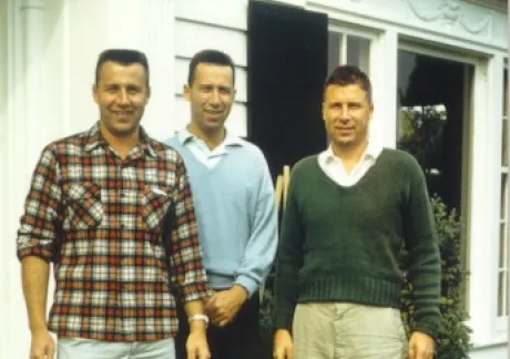 Three men standing in front of a white building with a brick step, dressed in casual clothing: one in plaid, one in a light blue sweater, and one in a green sweater.