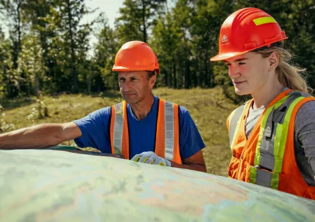 Two people wearing orange safety vests and hard hats look at a large map outdoors in a wooded area on a sunny day.