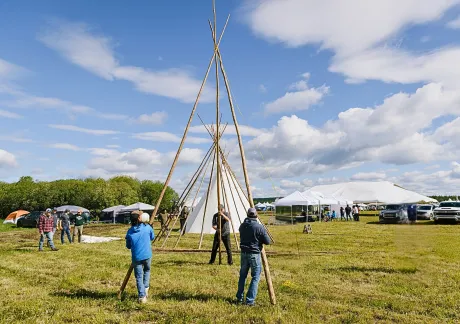 People are assembling a large teepee frame on a grassy field, with tents, parked vehicles, and cloudy blue skies in the background.