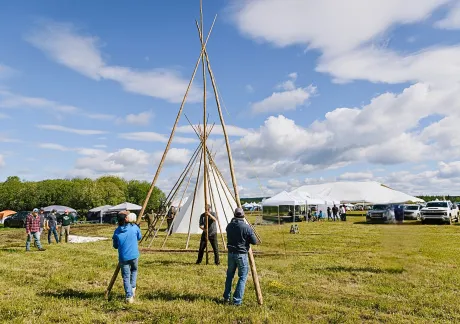 People are assembling a large teepee frame on a grassy field, with tents, parked vehicles, and cloudy blue skies in the background.