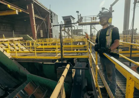 A worker wearing safety gear stands on a metal walkway in an industrial facility with machinery, railings, and equipment visible in the background.