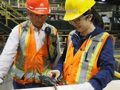 Two workers in safety gear, one in an orange helmet and the other in a yellow helmet, measure a piece of wood in an industrial setting.