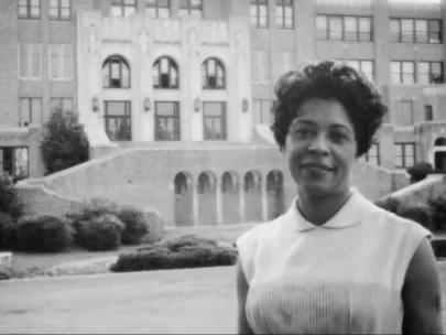 A woman is standing in front of a large building with multiple windows and a grand staircase. She is wearing a sleeveless shirt and smiling at the camera.