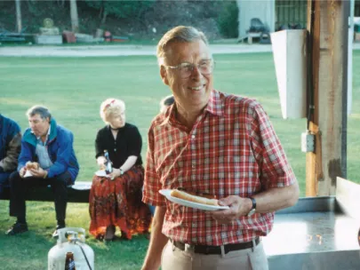 A man in a red checkered shirt holds a hot dog on a plate while standing outdoors at a social gathering. Three people are seated in the background.