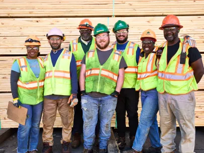 A group of seven construction workers wearing safety helmets and reflective vests stand together in front of a stack of wooden planks.