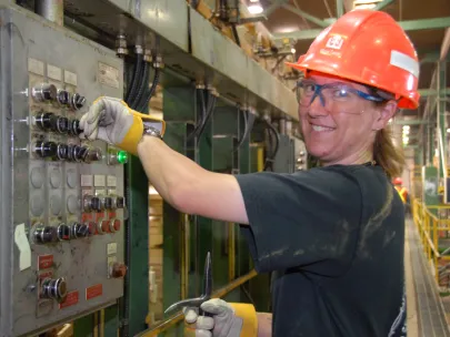 A person in a hard hat and safety glasses operates control buttons on an industrial panel, holding a wrench and smiling.