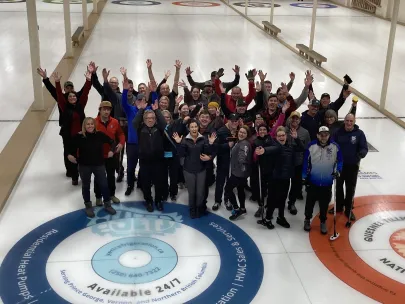 A group of people stand on an indoor curling rink, many with their arms raised, smiling at the camera.