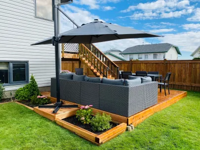 A backyard patio featuring a sectional sofa, umbrella, dining set, and small garden bed. The area is enclosed by a wooden fence and adjacent to a house with wooden stairs.
