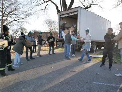 Volunteers work together to unload and distribute boxes of supplies from a truck parked on a street. Some volunteers form a line to pass the boxes down efficiently. Trees are visible in the background.