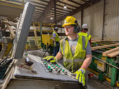 Worker in a safety helmet and high-visibility vest operates a control panel in an industrial setting, with other workers in the background.